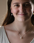 A smiling young woman with long brown hair wearing a delicate silver chain necklace with a small white pearl pendant and matching drop earrings, shown in a close-up portrait.