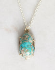 A silver chain necklace with an oval turquoise pendant set in a gold-toned prong setting, resting on a light marble surface.