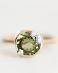 A close-up shot of a ring featuring a large oval green gemstone in a gold setting, accented by three small round clear stones on the upper right, against a plain light background.