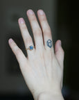 A close-up of a person's hand wearing a gold ring with a round blue gemstone, set against a dark, blurred background.