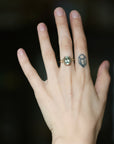 A close-up of a person's hand wearing a ring with a large oval light green gemstone in a gold setting, with a small geometric tattoo on the adjacent finger against a dark blurred background.