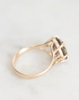 A close-up shot of a gold ring with a basketweave setting holding a smoky, translucent gemstone, resting on a light marble surface.
