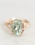 An oval-cut light green gemstone ring set in a rose gold prong setting, shown on a soft, light gray background.