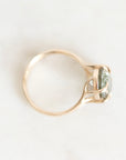 A gold ring with a twisted band and a bezel-set light green gemstone, photographed from above on a light off-white background.