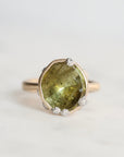 A gold ring featuring a large, irregular-cut olive green gemstone in a bezel setting, accented with small barnacles, photographed on a light gray background.