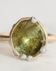 A gold ring featuring a large, irregular-cut olive green gemstone in a bezel setting, accented with small barnacles, photographed on a light gray background.