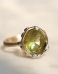 A silver ring featuring a large, faceted olive-green gemstone in a prong setting, with small barnacles along the band, photographed on a light beige background.