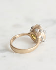 A gold ring with a textured, organic-shaped setting adorned with small barnacles, photographed on a light marble surface.
