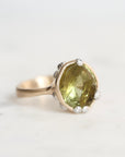 A vintage-style ring featuring a large, irregular-cut olive green gemstone in a gold-toned setting, accented with barnacles around the perimeter, photographed on a light marble surface.