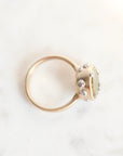 A gold ring with a round bezel-set pale green stone and small clear accent stones, photographed on a light marble surface.