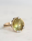 A gold ring featuring a large, faceted olive-green gemstone surrounded by small clear accent stones, set against a soft, light gray background.