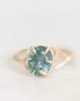 A gold ring featuring an oval-cut light blue gemstone in a prong setting, shown against a soft, light gray background.