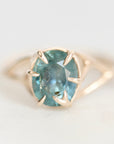 A gold ring featuring an oval-cut light blue gemstone in a prong setting, shown against a soft, light gray background.