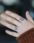 A close-up of a person's hand wearing a gold ring with a round light blue-green stone, set against a dark, blurred background. The person is wearing a brown knit sweater.