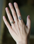 A close-up of a hand wearing a gold ring with a round clear gemstone, set against a softly blurred dark background.