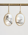 A pair of gold-tone oval drop earrings with mother-of-pearl cabochons featuring black ink drawings of mermaids, hanging from a gold rod against a light gray background.