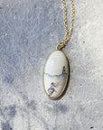 An oval pendant necklace with a gold-toned frame and chain, featuring a hand-painted scenic illustration of a lighthouse and a woman on a white background, resting on a textured gray stone surface.