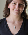 A woman with brown hair wearing a black V-neck top and a delicate gold chain necklace with a small white oval pendant, photographed in a softly lit indoor setting.