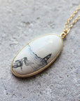 An oval pendant necklace with a gold-tone border and chain, featuring a cream-colored stone or resin inlay printed with a black ink-style landscape of a lighthouse and trees, resting on a textured gray stone surface.