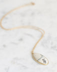 A gold chain necklace with an oval pendant featuring a white stone with black and blue veining, displayed on a light gray marble surface.