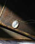 A close-up of a vintage oval pendant necklace with a white and gray bird design set in gold, resting on an antique brown leather-bound book.