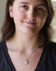 A woman with brown hair wearing a delicate gold chain necklace with a small oval white shell pendant, photographed in a close-up portrait.