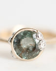 A close-up shot of a ring featuring a round, faceted green gemstone in a silver-toned setting, accented with three small clear crystals on the right side, against a plain white background.