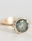 A close-up shot of a vintage-style ring featuring a large, round, pale green-gray gemstone in a gold bezel setting, accented by small clear stones on the shoulders, set against a soft, light gray background.