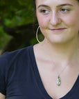 A woman wearing a silver-toned necklace with a small oval pendant and matching large hoop earrings, photographed outdoors with a blurred green background.