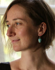 A woman in profile wearing a small turquoise drop earring, photographed indoors with soft natural light from a window behind her.