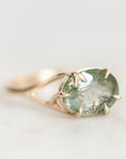 A gold ring featuring an oval-cut light green gemstone in a prong setting, shown on a soft, light gray background.