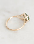 A rose gold ring with a square-cut green gemstone in a prong setting, resting on a light marble surface.