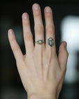 A close-up of a person's hand showing two rings: a small gold ring with a green oval stone on the ring finger, and a black geometric line tattoo on the middle finger. The background is dark and blurred.