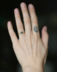 A close-up of a person's hand wearing a delicate gold ring with a small green gemstone, set against a dark, blurred background.