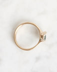 A delicate gold ring with a small marquise-cut green gemstone and a tiny clear accent stone, photographed from above on a white marble surface.