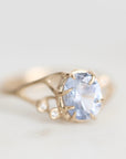 A gold ring featuring a large oval light blue gemstone in a prong setting, with small round accent stones along the band, photographed on a soft light gray background.
