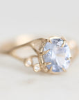 A gold ring featuring a large oval light blue gemstone in a prong setting, with small round accent stones along the band, photographed on a soft light gray background.