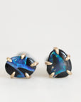 A pair of stud earrings featuring irregular dark blue and black opal stones in gold prong settings, displayed on a clean white background.