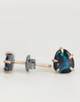 A pair of stud earrings featuring a dark, iridescent blue-green opal stone in a gold prong setting, shown on a plain light background.