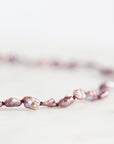 A close-up of a beaded necklace made of irregular, lustrous baroque pearls in a dusty lavender-purple shade, strung on a dark cord against a bright, soft-focus white background.