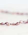 A close-up of a delicate necklace featuring irregular, baroque-style pinkish-purple freshwater pearls strung on a dark cord, resting on a light, neutral surface.