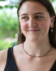A woman with brown hair wearing a delicate beaded necklace with pink, purple, and white pearls, along with small gold hoop earrings, set against a soft-focus green outdoor background.