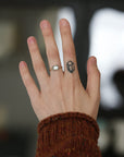 A close-up of a person's hand wearing two rings: a silver band with a small white oval stone on the ring finger, and a black geometric tattoo or stamp on the middle finger. The hand is wearing a brown knit sweater cuff, with a blurred dark background.