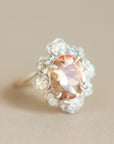 A close-up shot of a vintage-style ring featuring a peach-pink oval center stone surrounded by a halo of small clear stones and a gold band, set against a solid beige background.