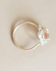 A delicate ring with a thin gold band, a silver-toned filigree setting, and a central oval pinkish-red gemstone, photographed on a neutral beige background.