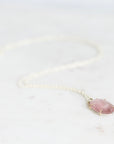 A delicate white beaded chain necklace with a small, oval-shaped pink gemstone pendant in a thin silver bezel setting, resting on a light marble surface.