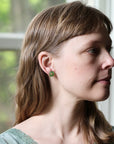 A woman in profile wearing a small green flower-shaped stud earring, photographed indoors near a window.