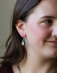 A close-up of a woman wearing a teardrop-shaped dangle earring with a pale green stone and dark blue accents, shown against a plain beige background.