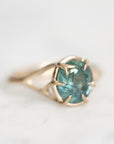 A gold ring with a round teal-blue gemstone in a prong setting, photographed close-up on a light marble-like surface.