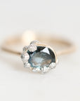 A close-up shot of a ring featuring a large oval blue-gray gemstone in a silver-toned setting, accented with small clear stones, on a soft white background.